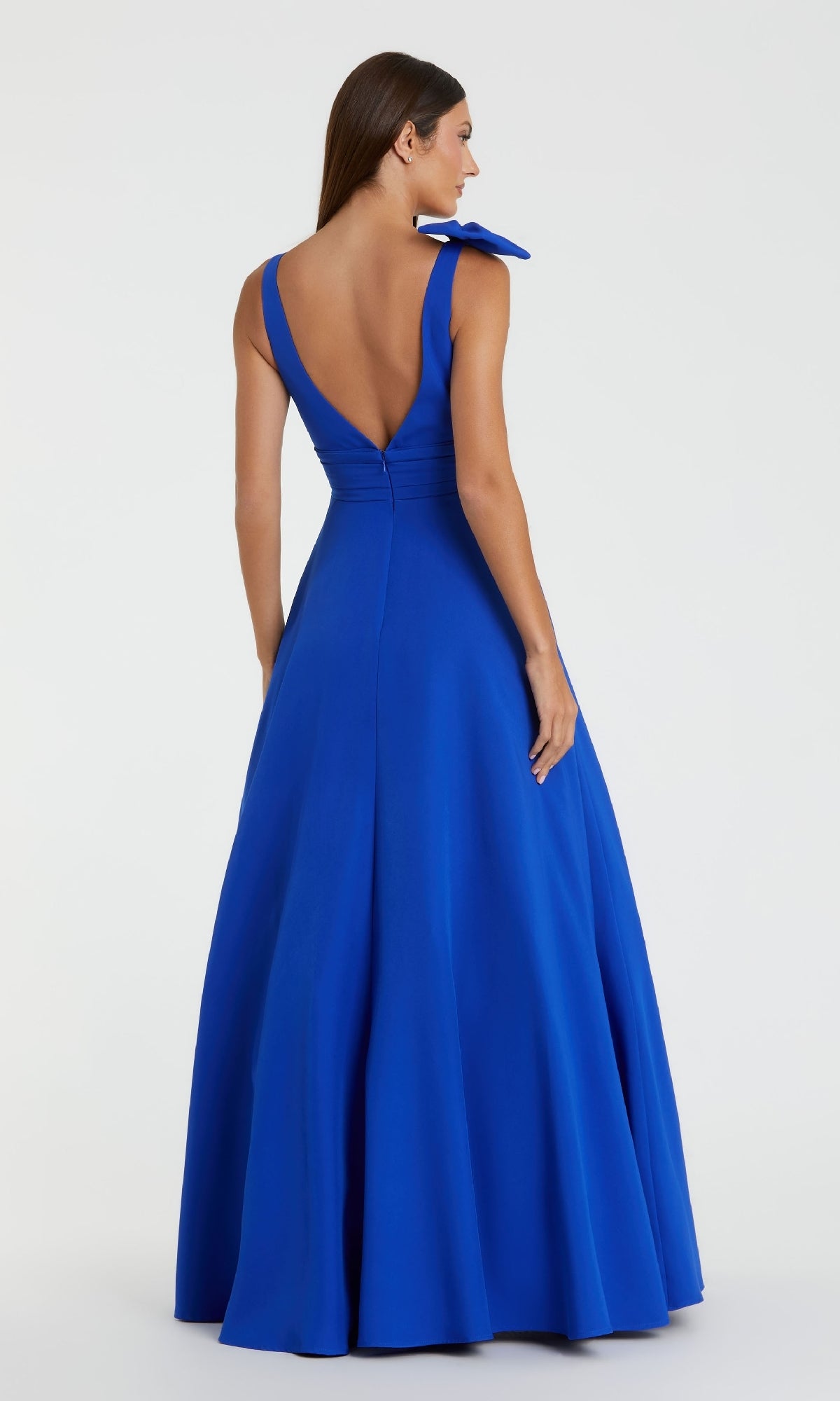 A woman in the Mac Duggal 49972 Shoulder-Bow Long Formal Dress, a floor-length royal blue gown with a sleeveless cut, deep V-back, and elegant shoulder bow detail, stands facing away on a plain light background.